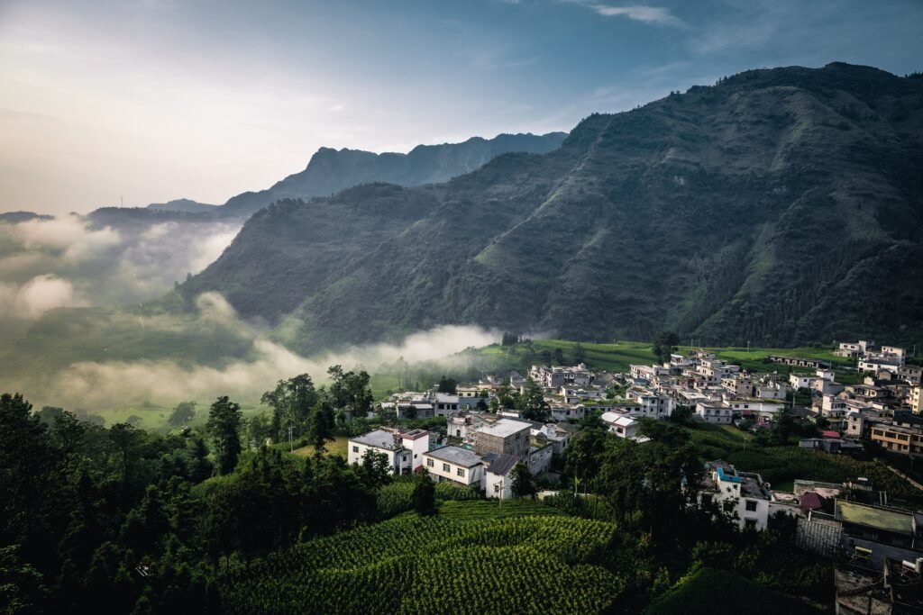 A serene village nestled in lush green mountains with morning mist drifting over the landscape.